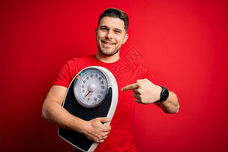 蓝眼睛的年轻健身男子在红色背景下拿着体重秤减肥图片