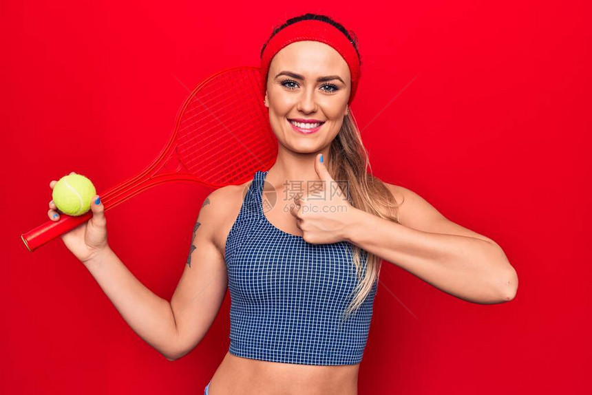 玩网球的年轻金发美女女员拿着拍打和球在红色背景上微笑快乐而积极拇指举起手表现优异图片