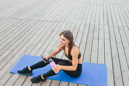 穿着运动服坐在瑜伽席子上在智能手机室外木板上聊天的图片