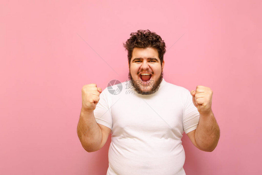 一个穿着白色T恤和超重的快乐男人的肖像为他心爱的球队在粉红色背景下的胜利而欢欣鼓舞快乐的留着胡子的胖开朗的男人高兴地举起双图片