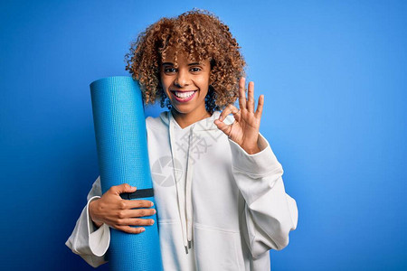做运动的年轻非洲裔美国女在孤立的蓝色背景上拿着瑜伽垫图片