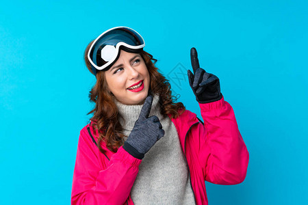 穿着滑雪眼镜的滑雪女人在孤立的蓝墙上用食指图片