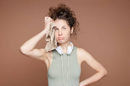 疲倦的女运动员从疲劳中叹气转动眼睛用毛巾擦掉前额汗水积极进行心肺训练在脖子上戴耳机和短须卡其顶部背景图片