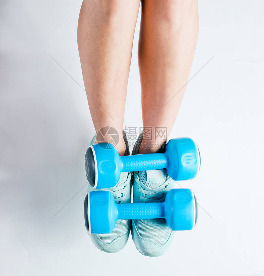 穿着运动鞋的女腿在白色背景上拿着蓝色塑料哑铃顶端视图适图片
