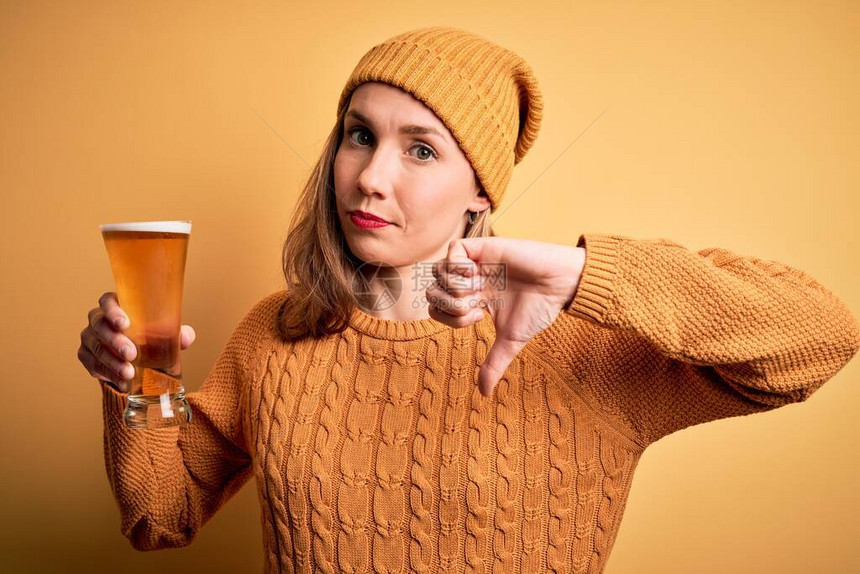 年轻美丽的金发美女在孤立的黄色背景上喝杯啤酒面脸愤怒图片