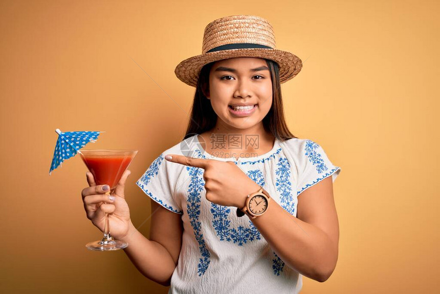 度假时戴着夏帽在黄色背景下喝鸡尾酒的亚洲小女孩非常高兴地用图片