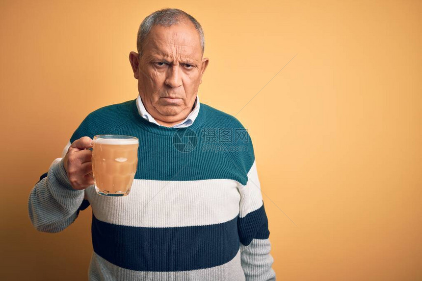 英俊的老人喝着一瓶啤酒站在孤立的黄色背景上怀疑和紧张图片