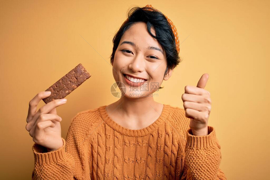 年轻美丽的亚洲女孩拿着能量蛋白质棒的麦片在孤立的黄色背景上快乐地微笑着做好牌子图片