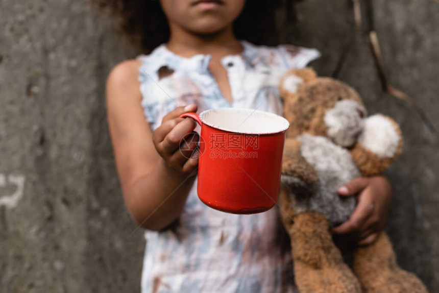 在城市街头乞讨时持有金属杯和泰迪熊的非洲贫困儿童被切图片
