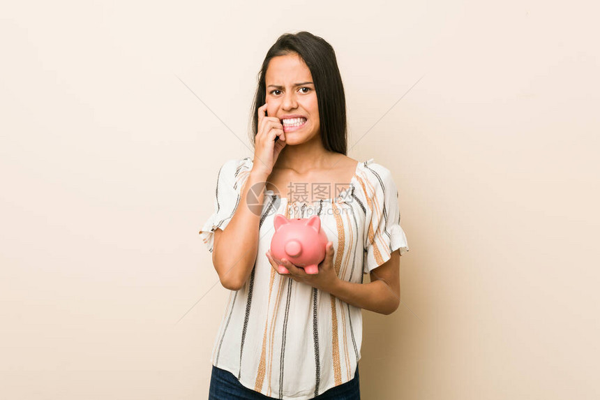 年轻的西班牙女人拿着小猪银行咬指甲紧张和非图片