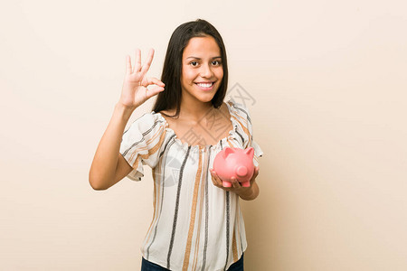 年轻的西班牙女人拿着小猪银行的快乐和自信表现出图片