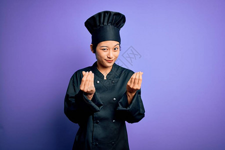 年轻漂亮的厨师女人穿着炊具制服和紫色背景的帽子图片
