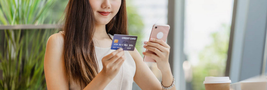 年轻的亚洲女使用带手机的信用卡在咖啡店或窗镜旁的合作空间进行网上购物技术货币钱包和在线支付概念图片