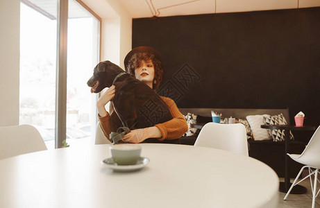 戴帽子的女士和狗坐在舒适的动物友好咖啡馆的桌子旁图片