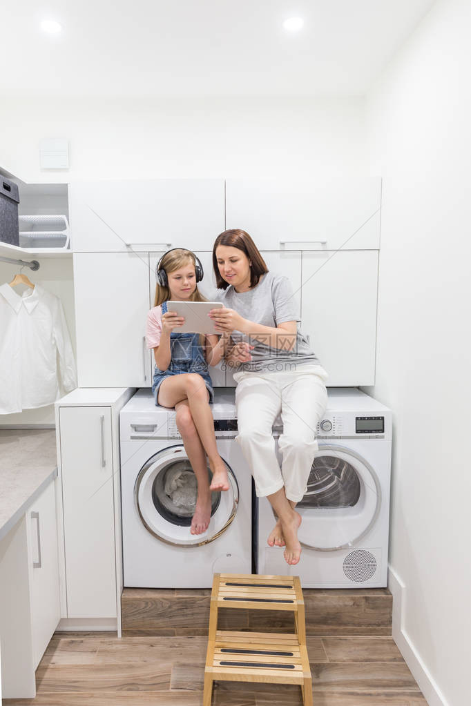 年轻的母亲和她的女儿正在洗衣房里坐在洗衣机图片