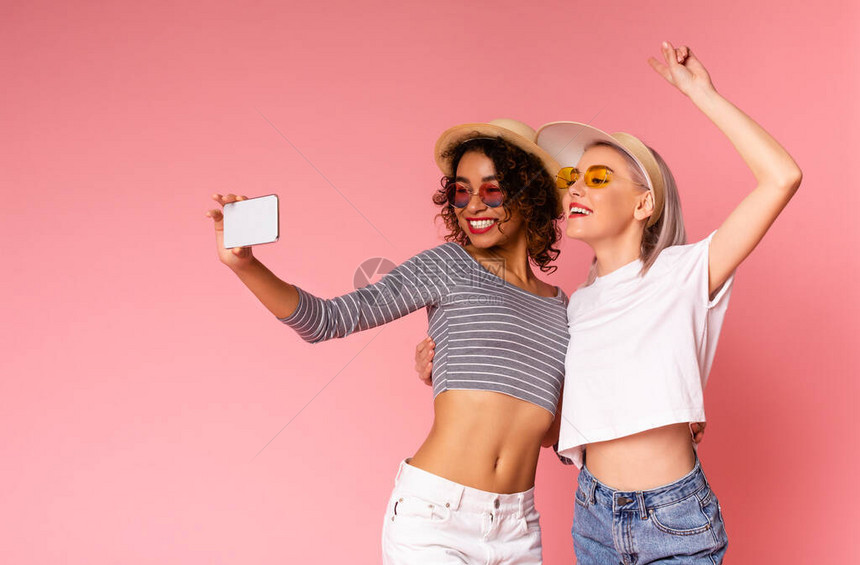 两个快乐的女朋友在夏季时档一起用智能手机自拍并展示和平姿态装成粉红图片