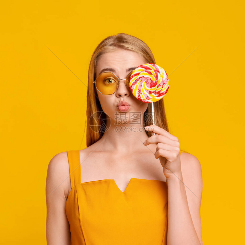 小甜点漂亮的年轻暑期女孩用大亮棒糖遮着眼睛看相机黄色背景和复图片