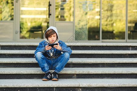 用智能手机在校外玩游戏的男孩技术网络成瘾和学校青少年在校舍前坐在楼图片