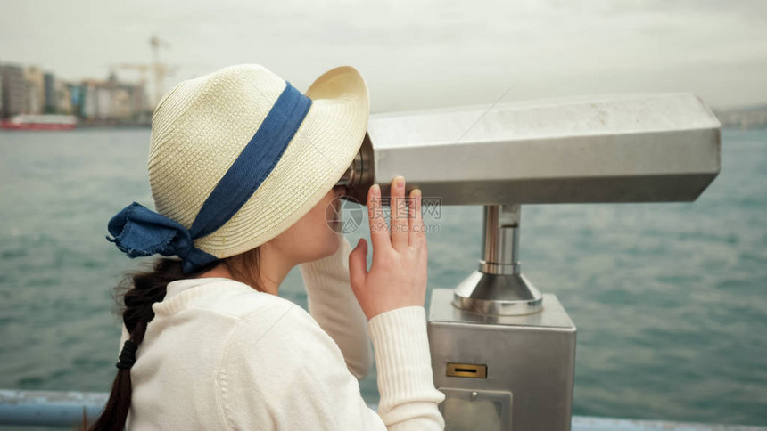 戴帽子的黑发女人透过双筒望远镜探索站在海边图片
