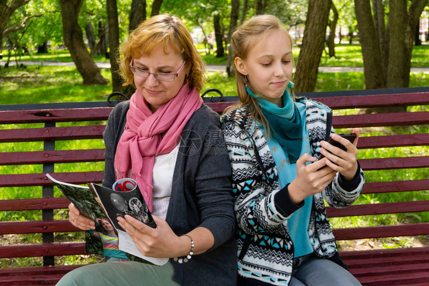 公园长椅上的未成年女儿和母亲正在阅读一些杂志的纸版和移动电子版本图片