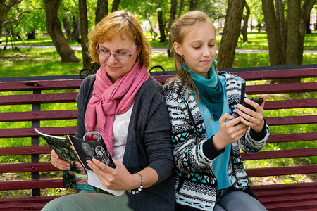 公园长椅上的未成年女儿和母亲正在阅读一些杂志的纸版和移动电子版本图片