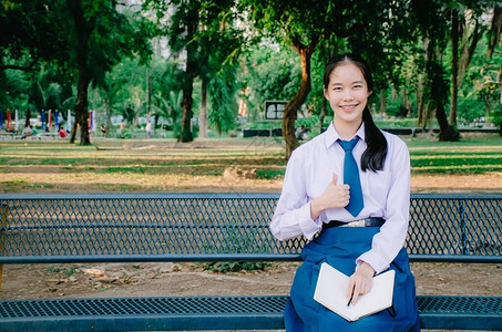 穿着校服阅读书和拇指的可爱少女坐在公园的长凳上图片
