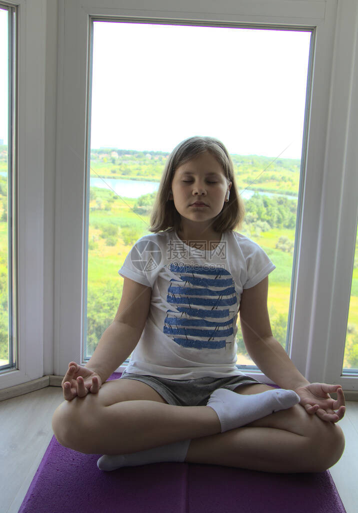 一个女孩正在窗边冥想孩子坐在地板上做瑜伽窗外的池塘和绿色草地上的景色大流行期间在图片