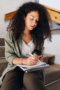 在大学楼梯上坐着深色卷发的年轻有吸引力的学生女孩背景图片