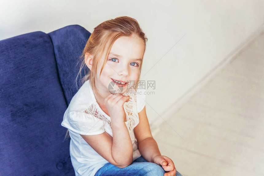 留在家里保持安全穿着牛仔裤和白色T恤的可爱小女孩坐在现代舒适的蓝色椅子上图片