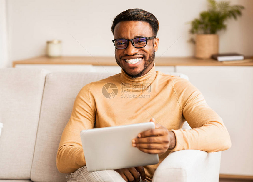 非洲裔美国戴眼镜持有数字石板和微笑在家中库奇的摄像机座椅上笑图片