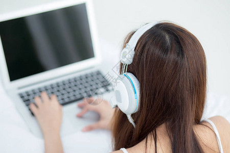 美丽的亚洲年轻女子在卧室度假时用耳机和笔记本电脑在线上网放松地听音乐图片