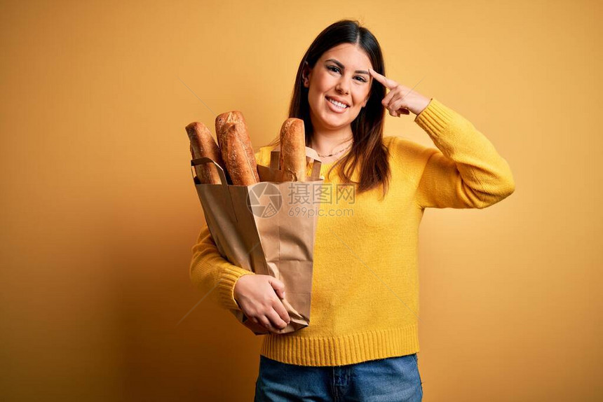 黄色背景上拿着一袋新鲜健康面包的年轻美女用一根手指微笑着指头图片