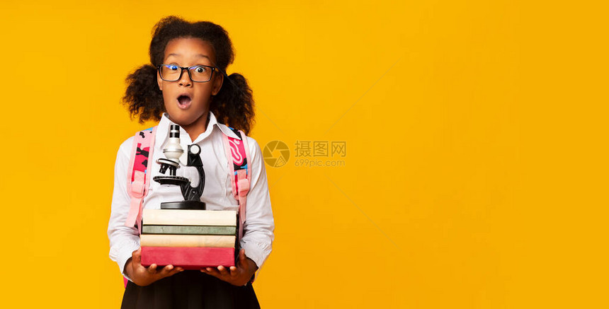 带着黄色工作室背景的显微镜和一堆书籍的惊吓黑人中学生女孩图片