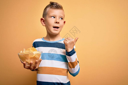 年轻的白人小孩在黄色背景上吃着不健康的土豆片薯图片