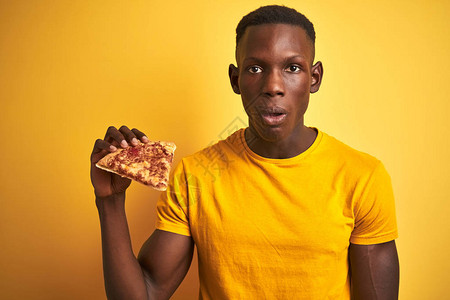 站在孤立的黄色背景上吃披萨的非洲裔美国人惊恐地惊讶地脸色惊恐图片