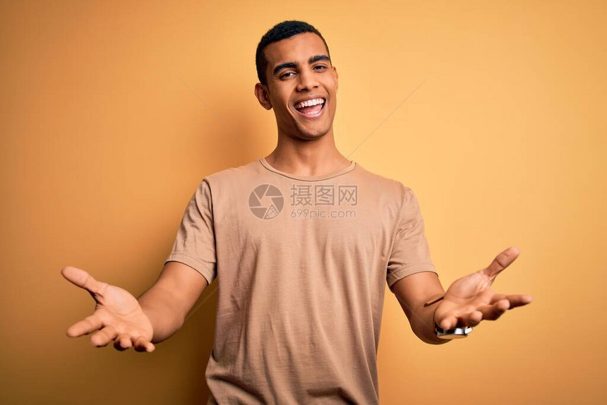 身穿轻便T恤的年轻英俊的非洲男子站在黄色背景上欢笑地微笑着伸出手来给予帮助和接受图片