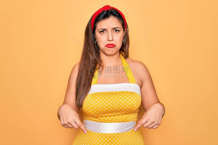 年轻的西班牙裔别针在黄色背景下穿着时尚感的50年代风格的女人指着向下看悲伤和不安图片