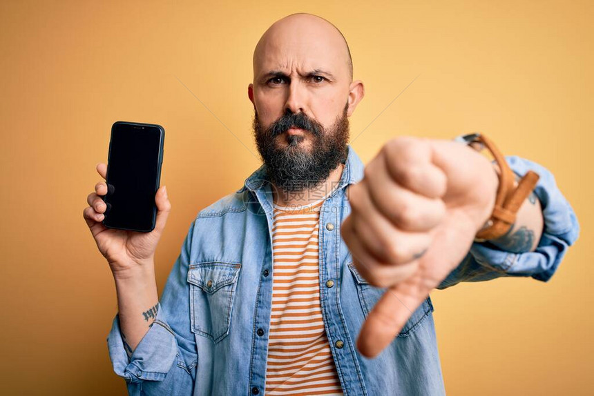 长胡子拿着智能手机的英俊光秃男子在黄色背景上用脸发怒反表情示不喜欢拇指低下拒绝概图片