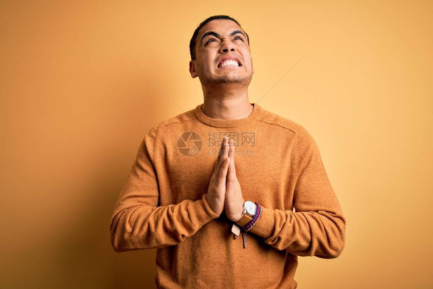 身穿散装毛衣站在孤立的黄色背景上乞讨和亲手祈祷与在脸上充满希望的表情的年轻男子图片