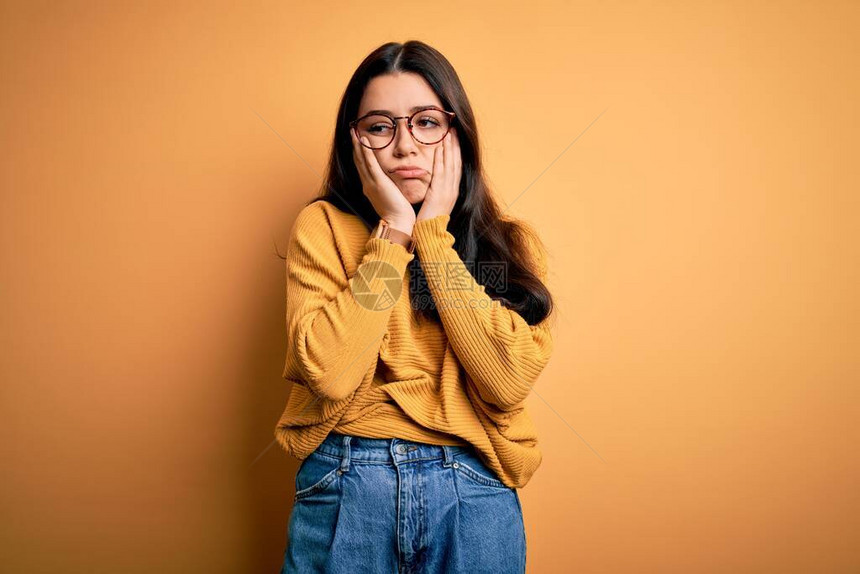 在黄色孤立的背景上戴着眼镜和休闲毛衣的褐发年轻女子疲惫的双手捂着脸抑郁和悲伤对问题感图片