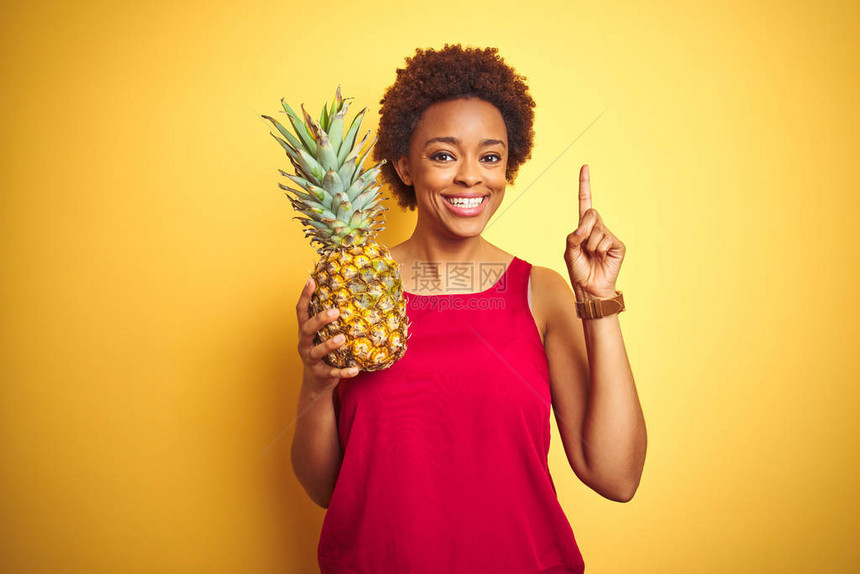 非洲裔美国妇女拿着热带菠萝在黄色孤立的背景下惊讶地发现一个想法或问题用快乐的脸指图片