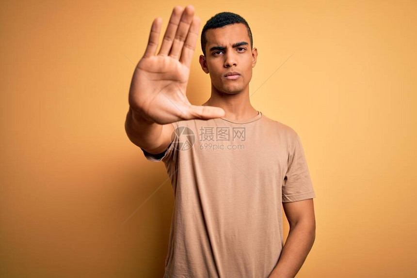 年轻英俊的非洲裔美国人穿着休闲t恤站在黄色背景上停止用手脸上带着消极和严肃的图片