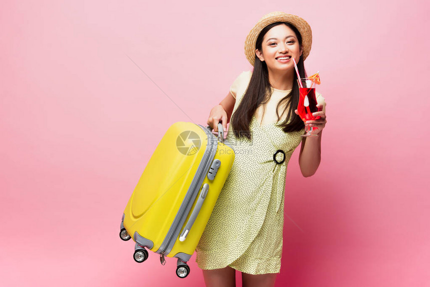 戴着草帽的微笑亚洲女孩拿着鸡尾酒和粉色的黄色行李图片