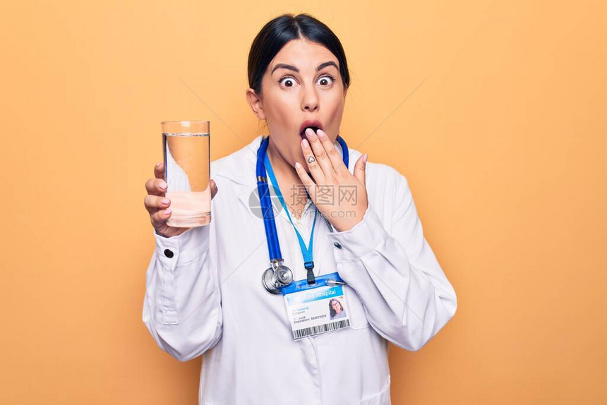 穿着听诊镜的美丽的女医生在黄色背景上喝水杯图片