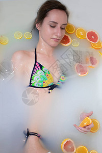 穿着鲜艳泳衣的女人在牛奶浴里放着橙子柠檬葡萄柚和书页健康水润的皮肤时装模特女孩水疗和护肤理念春天的颜色黄色背景图片