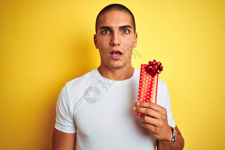 年轻的白人男子拿着生日礼物在黄色孤立的背景下惊恐地惊讶地脸色惊恐图片