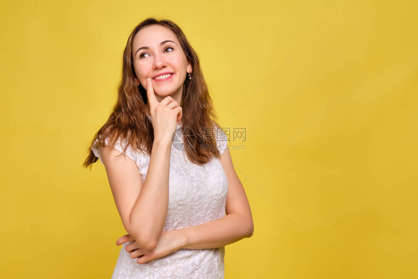 一个身穿白色T恤和黄色背景的棕色牛仔裤的女孩以梦幻般的外观愉快地思考着一图片