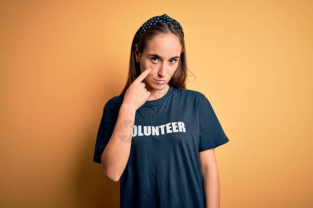 身穿志愿者T恤的年轻美女在黄色背景下做志愿者指着眼睛看着你的手势图片