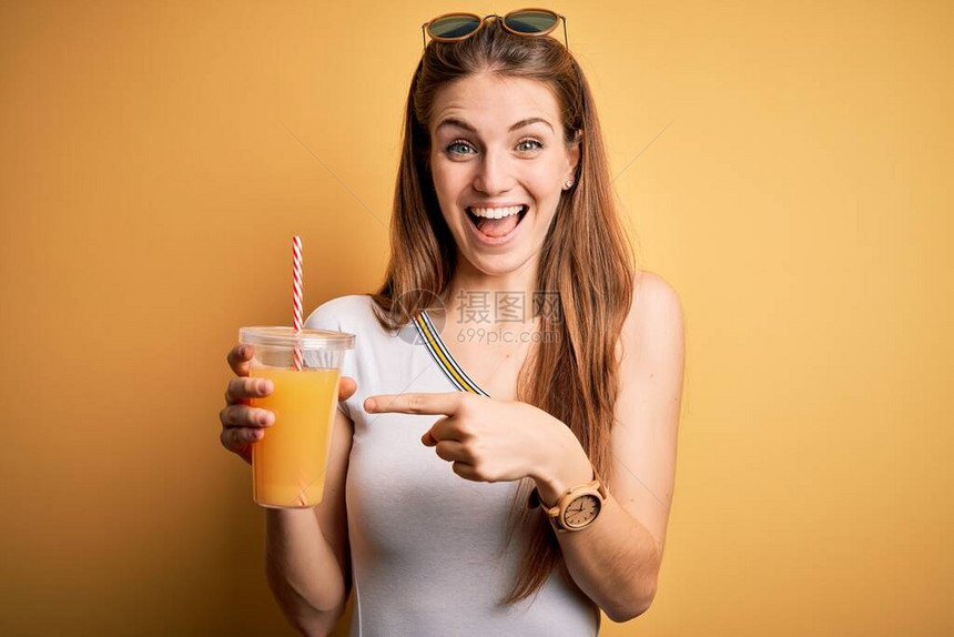 在黄色背景下喝健康橙汁的年轻漂亮红发女人非常高兴地用图片