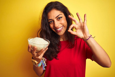 年轻美女拿着碗水和大米在孤立的黄色背景上边吃饭用手指做好标记图片